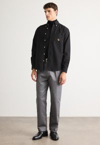 Hombre vestido con camisa negra de botones sobre un suéter negro de cuello alto, pantalones grises, cinturón negro y zapatos negros, de pie sobre un suelo gris contra una pared blanca.