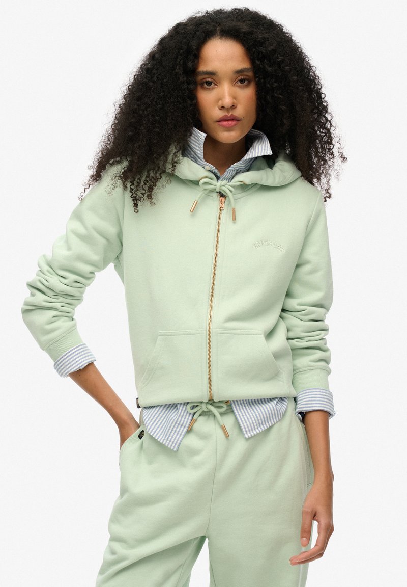 Felpa verde chiaro con zip, realizzata in tessuto morbido, dotata di cappuccio con coulisse, tasche frontali e dettagli in metallo dorato.