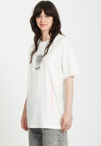 Camiseta blanca con un diseño gráfico de dos caras y el texto "VOLCOM" en el pecho. Presenta texto vertical rosa a lo largo de un lado. Mangas cortas.