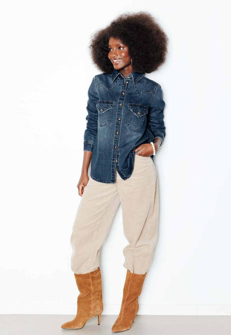 Femme aux cheveux afro portant une chemise en jean bleue, un pantalon en velours côtelé beige et des bottines en daim marron à talons, debout avec une main dans la poche.