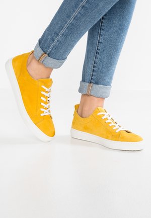 Gule ruskindssneakers med hvide snørebånd, båret med opkrogede blå jeans mod en ensartet hvid baggrund.