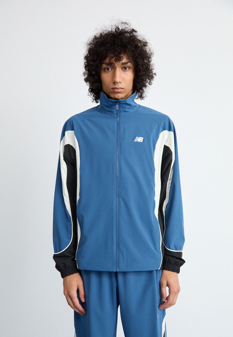Ung person med krøllet hår iført en blå, sort og hvid zip-up sportsjakke og matchende blå bukser, stående foran en ensfarvet baggrund.