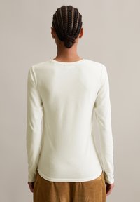 Eine Person mit geflochtenem Haar in einem niedrigen Dutt trägt ein weißes Langarmshirt und braune Hosen und steht mit dem Rücken zu einem schlichten Hintergrund.