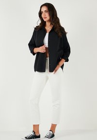 Camicia nera con bottoni, maniche lunghe, taglio rilassato, indossata sopra una maglietta corta bianca, abbinata a jeans bianchi alla caviglia e sneakers nere.