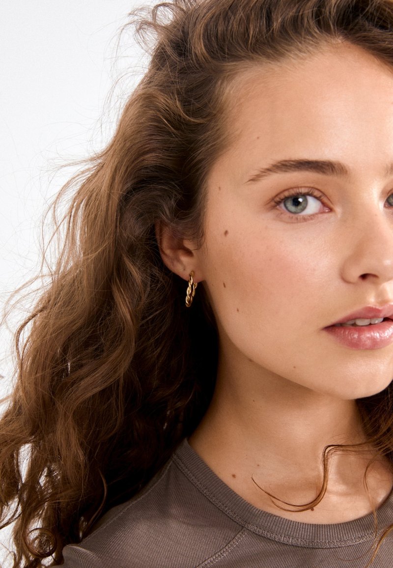 Gold metal hoop earring with a twisted design. Model has curly hair, wearing a fitted grey top. Natural makeup with neutral tones.