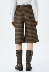 Culotte a quadri marroni con taglio dritto, caratterizzate da una vita visibile e passanti per la cintura. Abbinati a stivali alti in pelle marrone con fibbia.