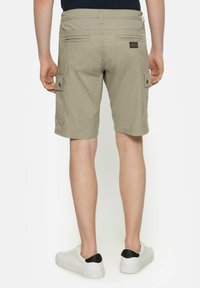 Mies, joka on pukeutunut vaaleanbeigeihin cargo-shorts-housuihin ja valkoisiin tennareihin, joissa on mustat kantapääkorostukset, seisoo yksinkertaisen valkoisen taustan edessä.
