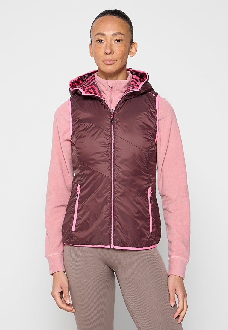 Frau trägt eine ärmellose burgunderfarbene Weste mit pinkem Besatz und zwei Reißverschlusstaschen über einem pinken Langarm-Fleece und beigen Leggings, steht vor einem schlichten Hintergrund.