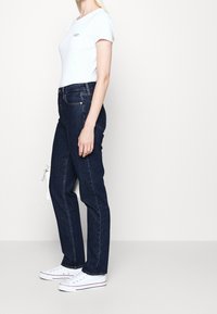 Woman standing sideways wearing a fitted white t-shirt, dark blue straight-leg jeans, and white sneakers against a plain background.