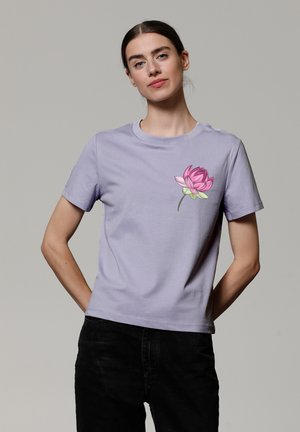 T-shirt lilas en coton avec des manches courtes, affichant un graphique de fleur de lotus rose sur le côté gauche de la poitrine. Design de col rond simple.
