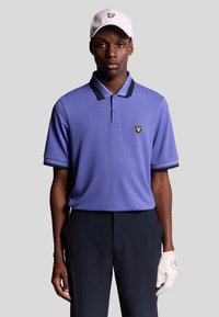 Uomo che indossa una polo viola chiaro con strisce nere sul colletto, pantaloni neri, guanto da golf bianco e cappellino bianco con logo di un uccello giallo.