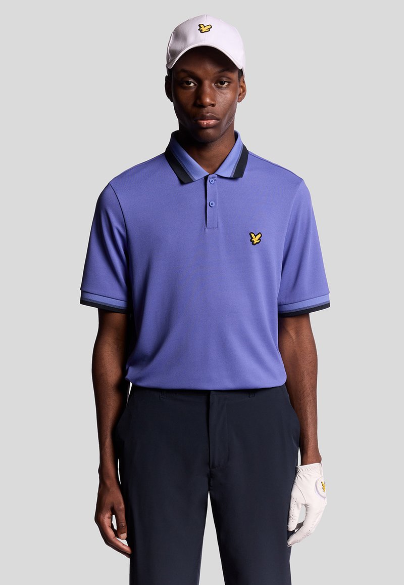 Uomo che indossa una polo viola chiaro con strisce nere sul colletto, pantaloni neri, guanto da golf bianco e cappellino bianco con logo di un uccello giallo.