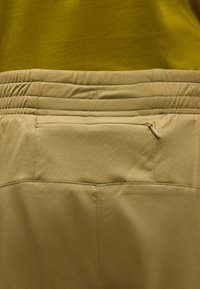 Κοντινό πλάνο ελαστικής μέσης σε μπεζ χρώμα με μια μικρή τσέπη με φερμουάρ σε ανοιχτό καφέ παντελόνι και μουσταρδί κίτρινο τοπ από πάνω.