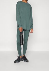 Långärmad grön topp kombinerad med matchande leggings. Svarta sportskor med texturerade detaljer. Användaren håller en svart sportflaska.
