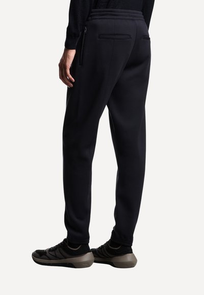 Schwarze Sporthose mit schmaler Passform, ausgestattet mit Reißverschluss-Rückentaschen und einem gerippten Bund. Kombiniert mit schwarz-grauen Sportschuhen.