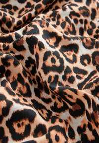 Látka s leopardím vzorem, která obsahuje výrazné černé skvrny na teplém oranžovém a smetakovém pozadí, prezentující měkký, strukturovaný povrch s volně spadajícími záhyby.