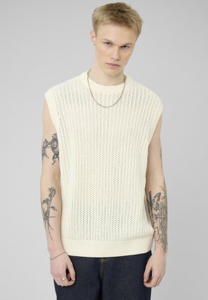 Jeune homme aux cheveux blonds courts portant un pull en maille blanche sans manches, un jean bleu, un collier chaîne et des tatouages visibles sur les bras.