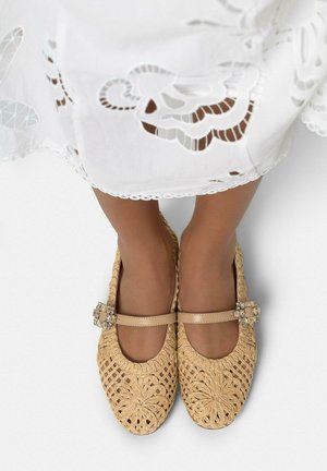 Pies calzados con zapatillas Mary Jane beige tejidas con hebillas adornadas, parcialmente cubiertos por una falda blanca bordada.