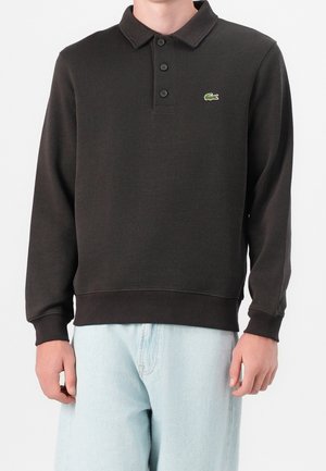 Homme portant un polo à manches longues marron foncé avec trois boutons et un petit logo crocodile vert sur la poitrine, associé à un jean bleu clair.