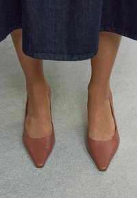 Scarpe con tacco alto in pelle marrone, con punta affilata e taglio laterale basso, indossate con una gonna in denim scura su una superficie grigia chiara.