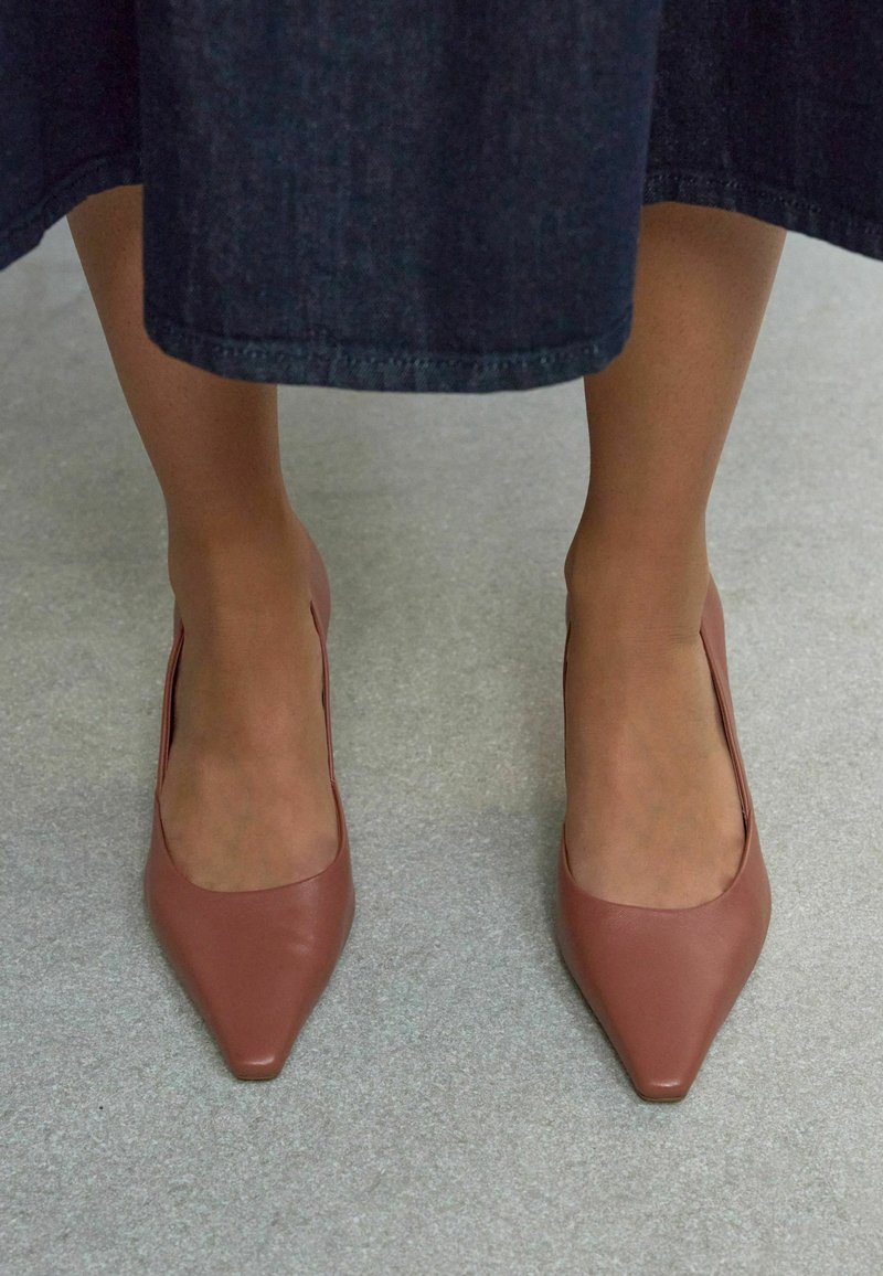 Scarpe con tacco alto in pelle marrone, con punta affilata e taglio laterale basso, indossate con una gonna in denim scura su una superficie grigia chiara.