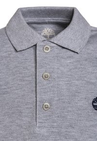 Szary polo z kołnierzykiem, trzema metalowymi guzikami i haftowanym logo. Wykonane z teksturowanej tkaniny z widocznymi detalami szwów.