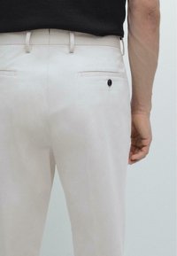 Lichtbeige broek met een gladde textuur, voorzien van achterzakken en een zwart knoopdetail. De tailleband is getailleerd met een ceintuurlus.