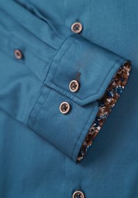 Tee in Türkis mit einer glatten Textur, verziert mit braunen Knöpfen und einem floral gemusterten Manschettenfutter in Blau- und Brauntönen.