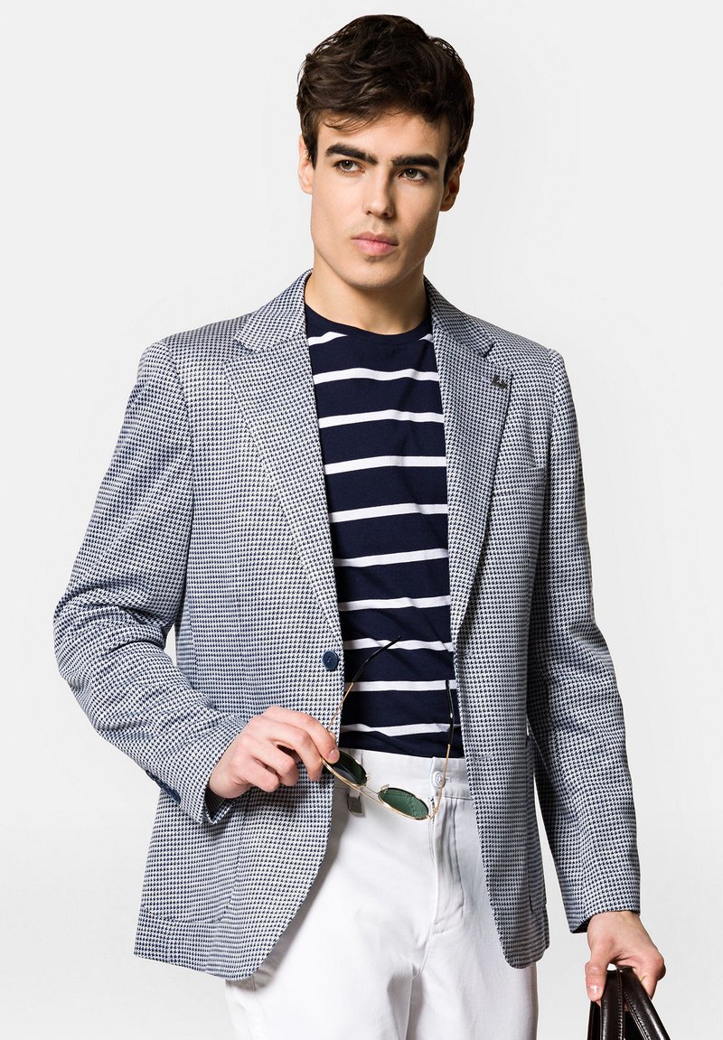 Marynarka w niebiesko-białą kratę houndstooth, z klapkami, na pojedynczy guzik, zestawiona z granatowym T-shirtem w paski i białymi spodniami.