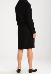 Schwarzer Wollmantel mit strukturiertem Kragen, tailliertem Design und Schlitz hinten, der bis zur Mitte der Waden reicht. Getragen mit schwarzen flachen Schuhen.