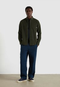 Camicia verde con bottoni, finitura testurizzata, taschino sul petto, abbinata a pantaloni blu navy e scarpe blu navy, in piedi su un pavimento grigio chiaro.