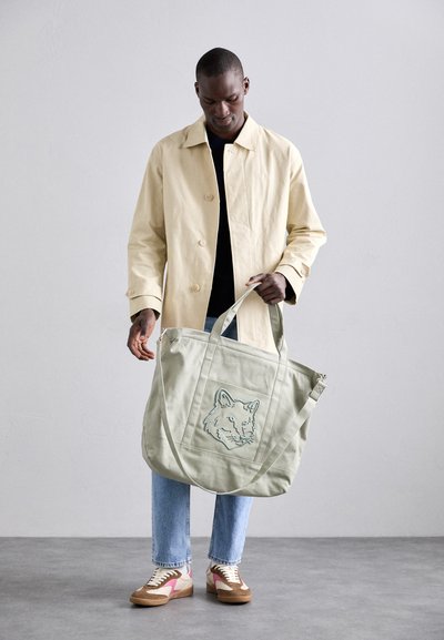 Bolsa de lona en verde claro con bordado de cara de lobo. El modelo también lleva una chaqueta beige, una camiseta negra y jeans de color azul claro.