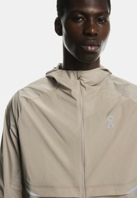 Gaiši brūns viegls mētelis ar rāvējslēdzēju, kapuci un atstarojošiem elementiem. Piedāvā logotipu uz krūtīm un gludu tekstūru.