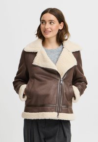 Veste en cuir marron avec col et poignets en shearling crème, fermeture éclair et poches latérales ; présente une texture lisse et une coupe ajustée.