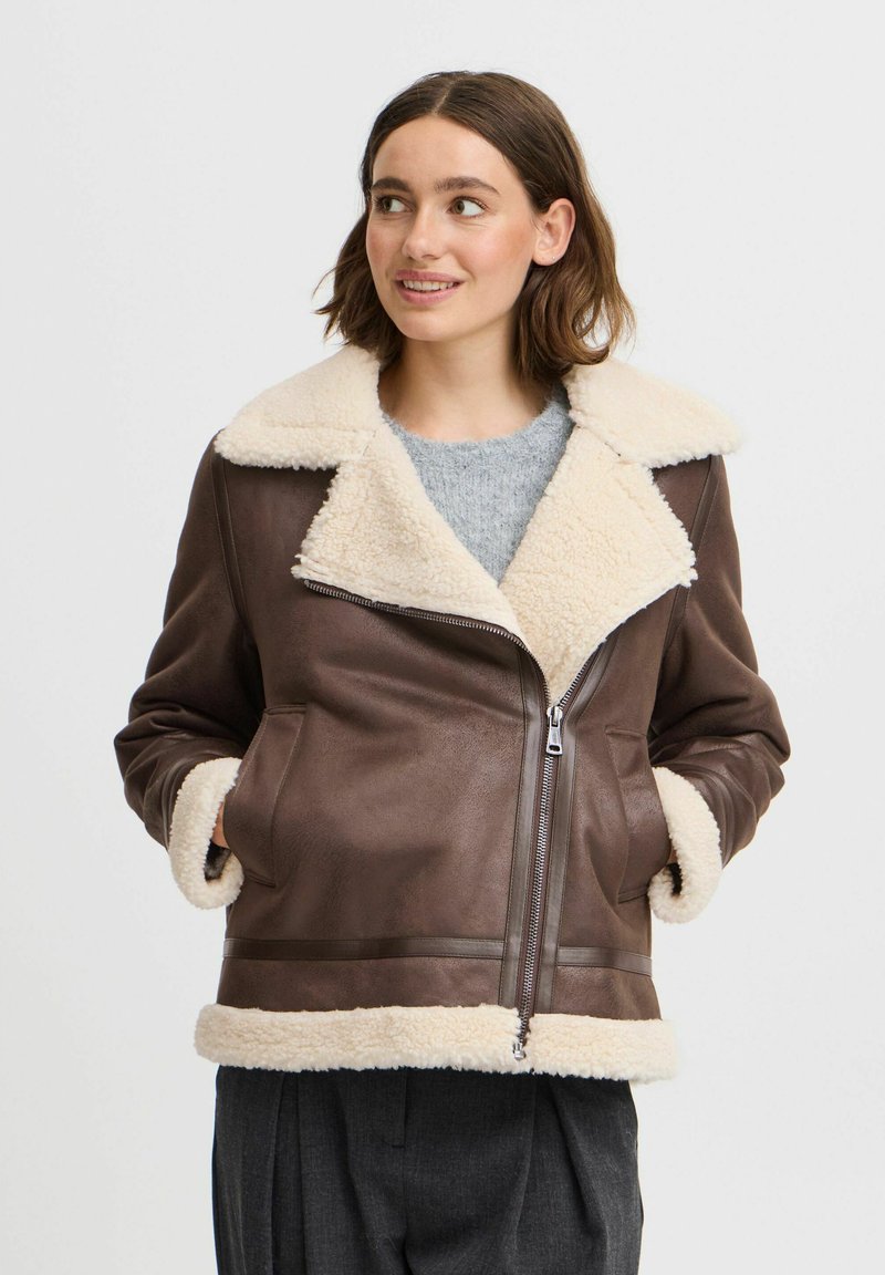 Veste en cuir marron avec col et poignets en shearling crème, fermeture éclair et poches latérales ; présente une texture lisse et une coupe ajustée.