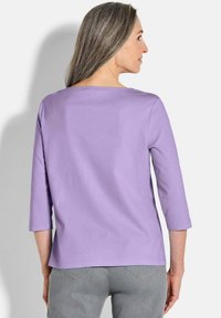 Helles lila Bluse mit Dreiviertelärmeln und großem Ausschnitt, aus glattem Material, getragen über grauen Hosen. Einfaches Design ohne Verzierungen.