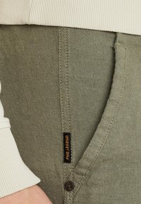 Pantalones jogger verde militar con una textura suave, cinturilla elástica, bolsillo lateral y etiqueta de marca negra cosida en la parte frontal.