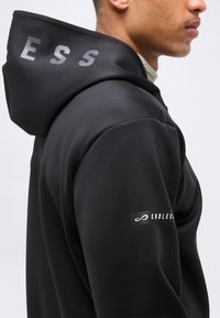 Homme portant un sweat à capuche noir avec le texte "ENDLESS" sur la manche et les lettres partielles "ESS" sur la capuche, montré de l'épaule vers le haut, vue de côté.