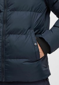 Mornarska puffer jakna s zatvaračem, rebrastim manžetama i bočnim džepovima, s laganim, quilted dizajnom i glatkom teksturom.
