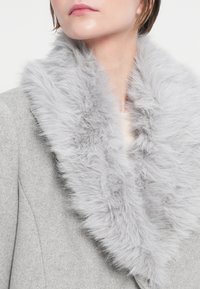 Abrigo de lana gris con un suave y esponjoso cuello de piel sintética gris, que resalta la textura y el calor. El cuello añade un detalle contrastante al abrigo.