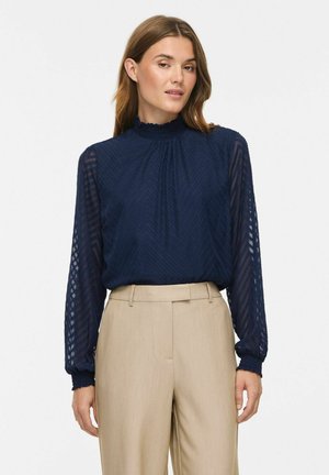 Femme portant un chemisier bleu marine à motifs avec de longues manches transparentes et un pantalon beige taille haute, debout devant un fond uni.