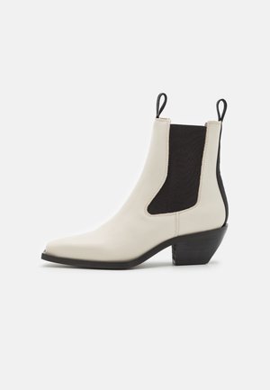 Bottine beige en cuir avec panneaux latéraux élastiques noirs, talon noir et languettes de tirage sur fond blanc.