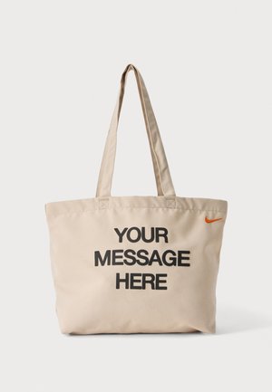 Beżowa torba na ramię z długimi uchwytami i czarnym napisem "YOUR MESSAGE HERE" nadrukowanym z przodu, z małym pomarańczowym logo Nike.