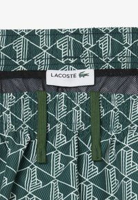 Groene shorts met een trekkoord, voorzien van een wit geometrisch patroon en een zwarte mesh voering, met een wit Lacoste-merketiket en groene trekkoorden.
