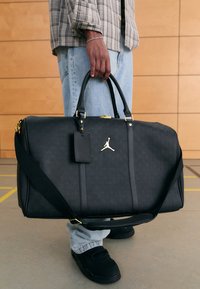 Svart duffelsekk laget av teksturert materiale med gullfarget beslag, "23"-mønster og justerbar rem. Har standard duffelstørrelse.