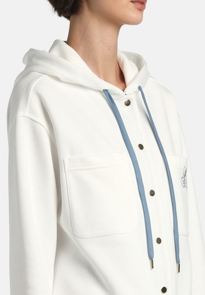 Prim-plan cu o persoană purtând o jachetă albă cu glugă, cu șnururi albastre, capse și două buzunare frontale la piept.