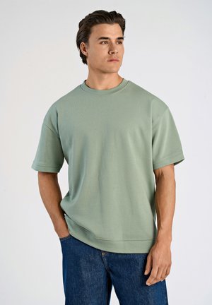 T-shirt oversize verde chiaro con maniche corte, colletto tondo e una texture liscia, abbinata a jeans di denim blu scuro.
