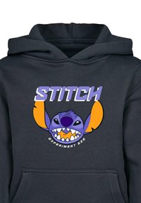 Čierna mikina s kapucňou so farebnou grafikou Stitcha, modrého mimozemšťana s oranžovými ušami a širokým úsmevom, text znie „STITCH“ a „EXPERIMENT 626.“