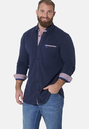 Bärtiger Mann, der lächelt, trägt ein marineblaues Hemd mit Knöpfen, gemustertem Kragen und Manschetten sowie hellblaue Jeans, Hand in der Tasche.