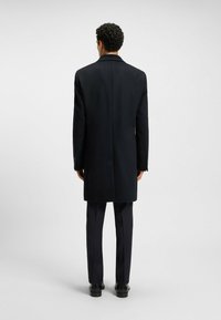 Cappotto di lana nera con taglio dritto, fessura singola e spalle strutturate. Abbinato a pantaloni scuri su misura e scarpe nere.